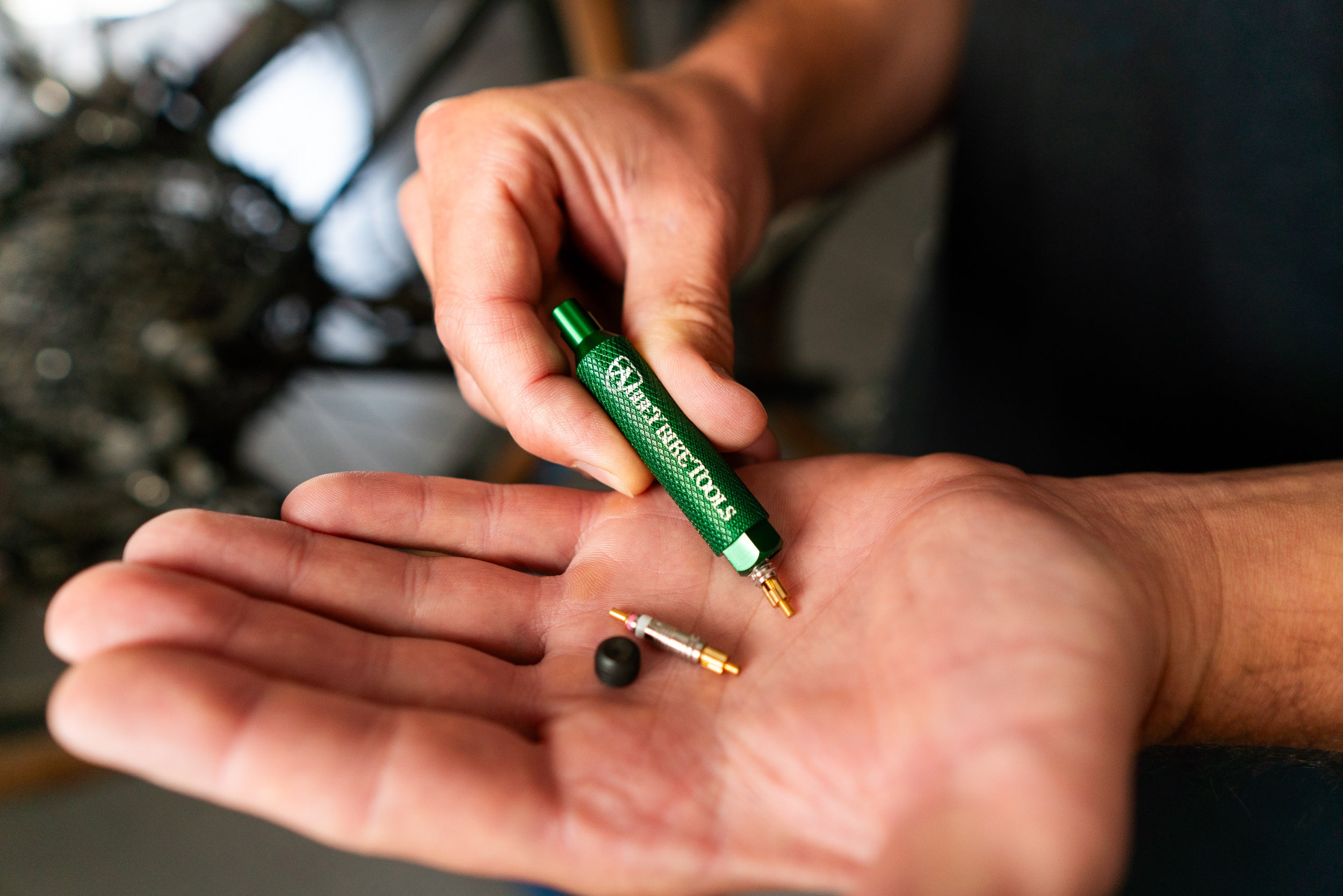 A close-up of a person's hand holding a green and gold bicycle valve stem tool over their other hand, which holds a removed bicycle tire inner tube and valve cap.