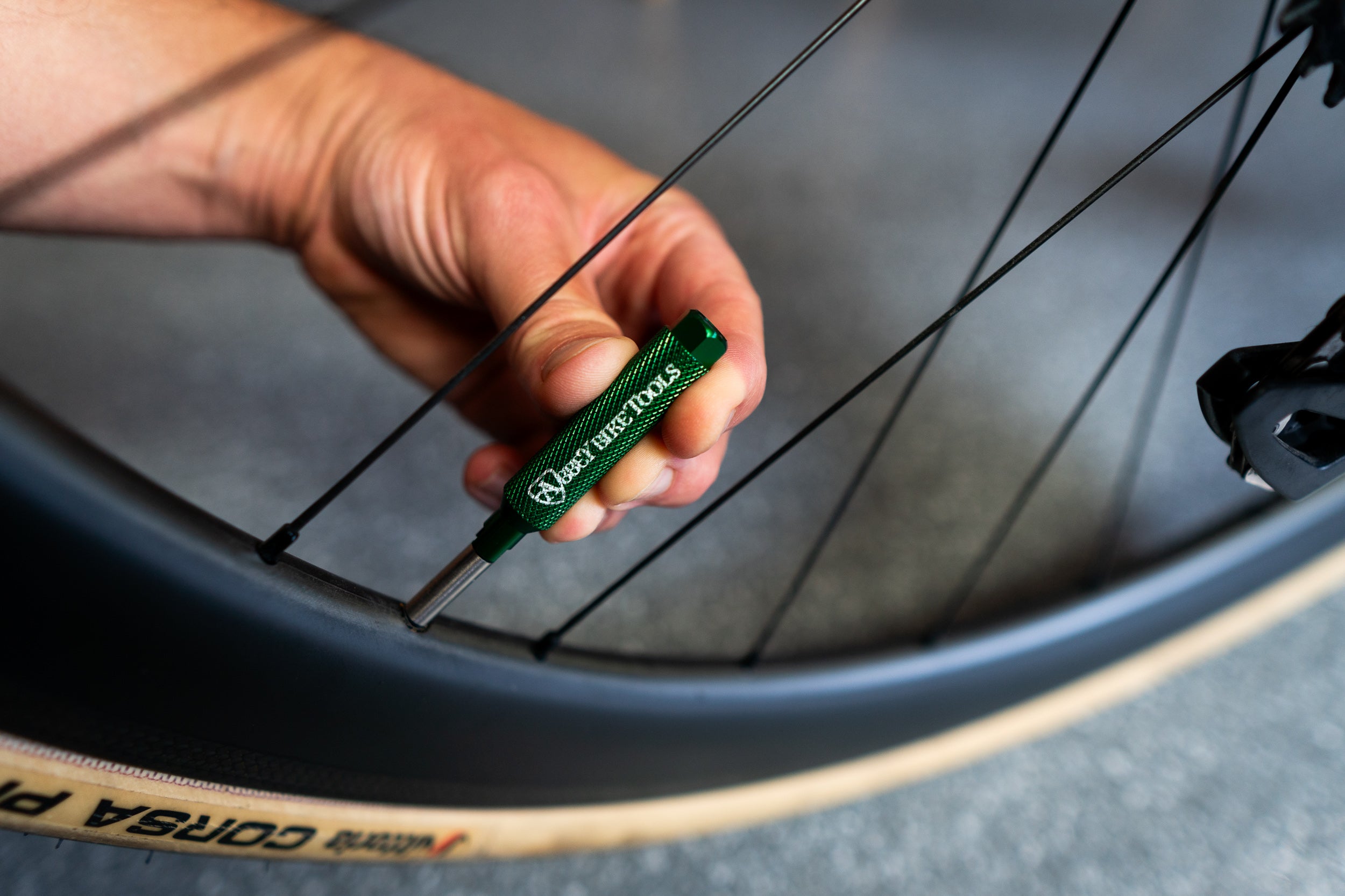 A close-up of a hand holding a green Presta Wrench tool against a bicycle wheel, with a Vitoria Corsa tire partially visible in the foreground.